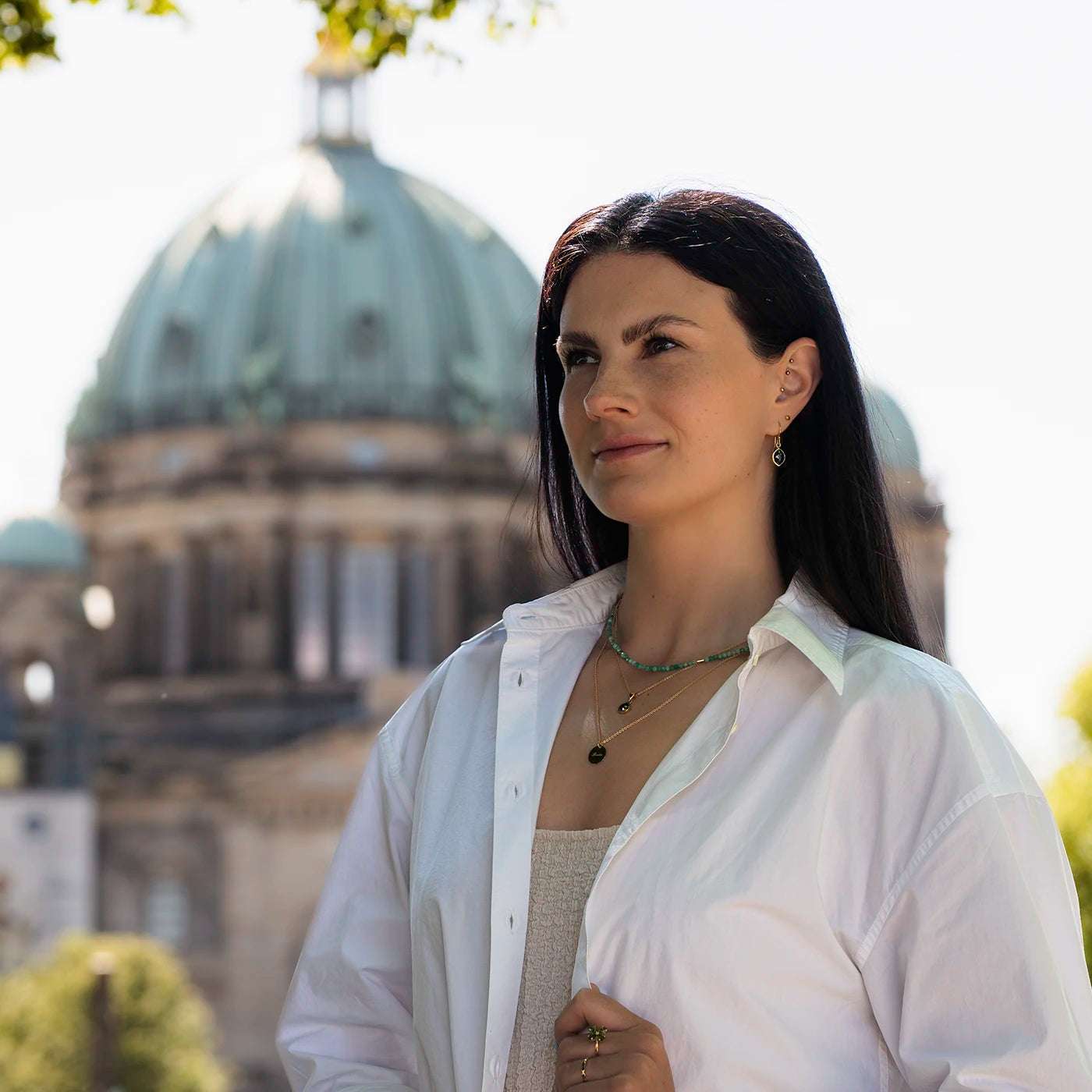 Gravurplaettchen rund Berlin Forever 0.5mm dick und 14mm Durchmesser Gelbgold mit Gravur Berlin an modelhals in Layering mit schwarzer Diamantkette und Edelsteinkette mit Smaragden halb nah vor Berliner Dom von unten schraeg