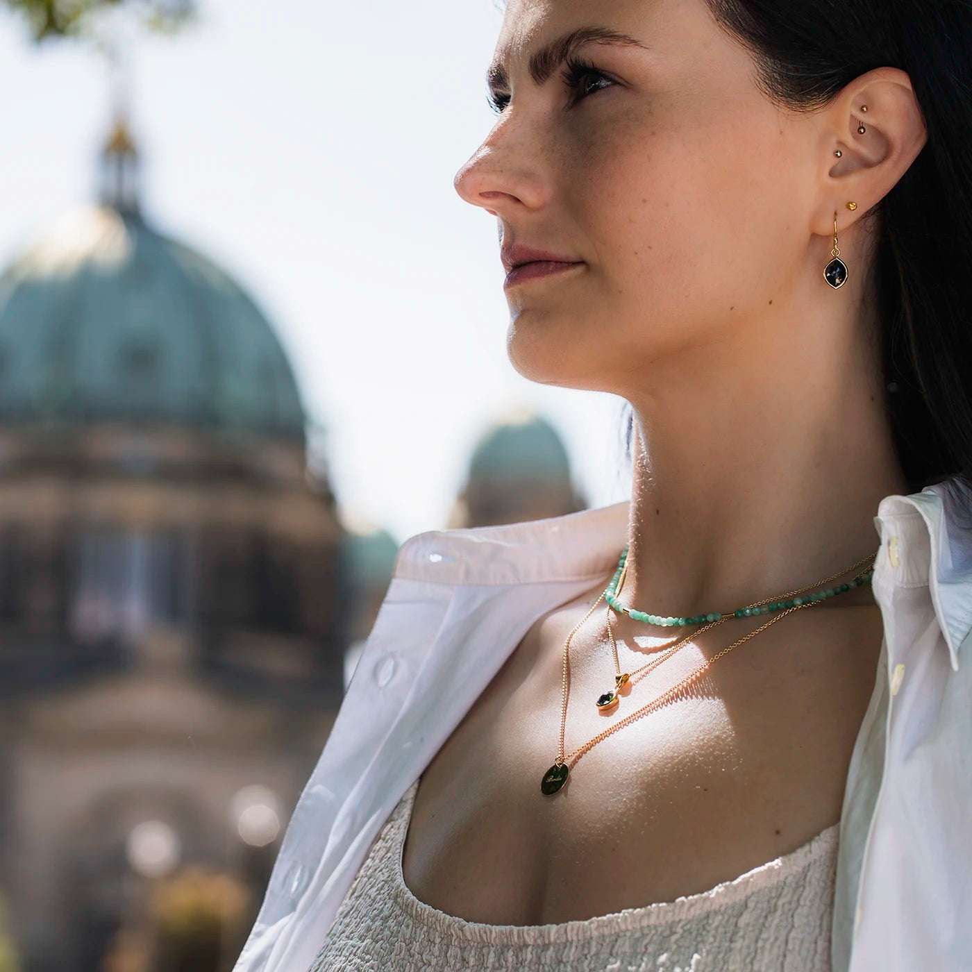 Gravurplaettchen rund Berlin Forever 0.5mm dick und 14mm Durchmesser Gelbgold mit Gravur Berlin an modelhals in Layering mit schwarzer Diamantkette und Edelsteinkette mit Smaragden halb nah vor Berliner Dom profilig