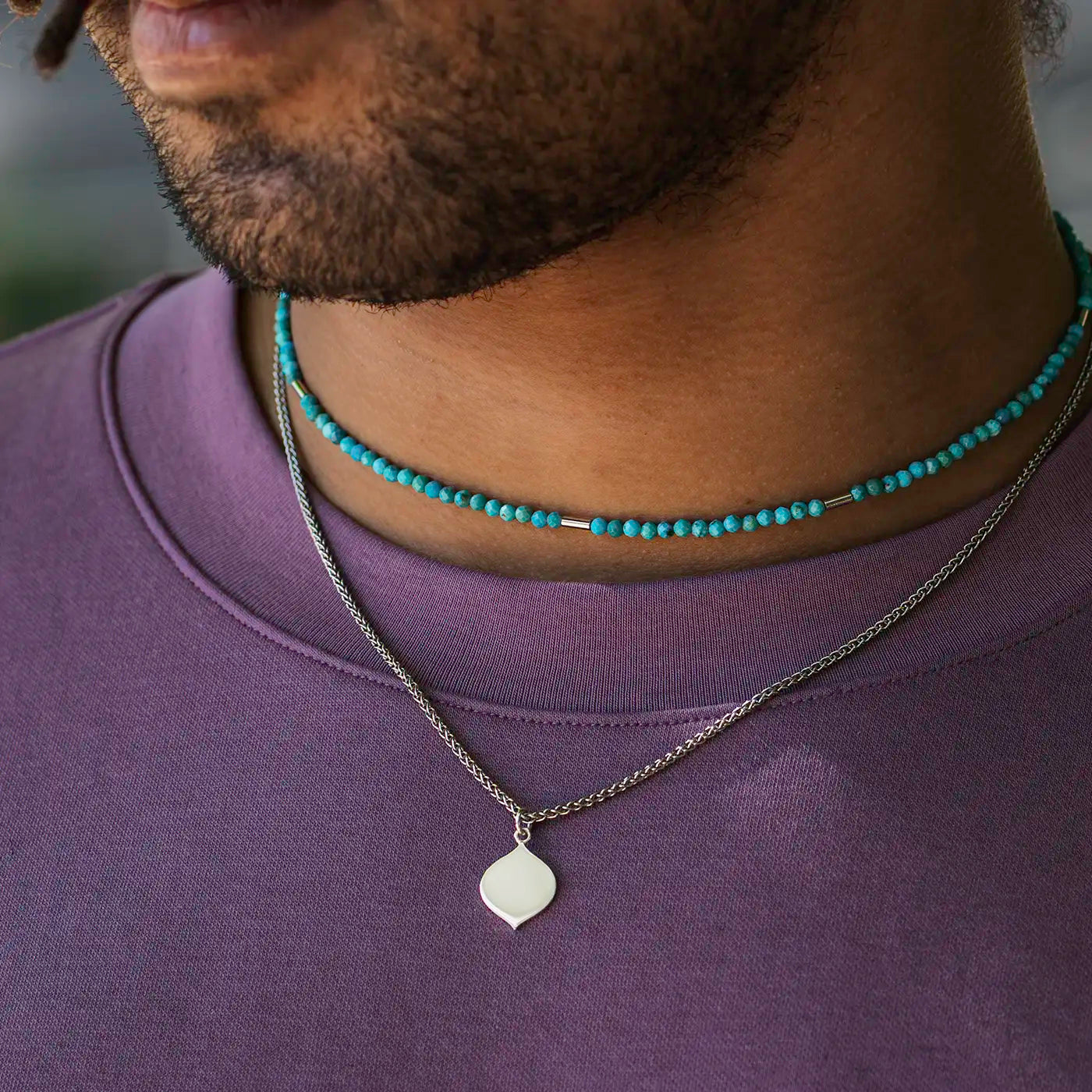 Eine Person mit Bart trägt ein lila Oberteil. Sichtbar sind zwei Halsketten: eine aus kleinen türkisfarbenen Perlen, die andere ist eine silberfarbene Kette mit einem ovalen oder rautenförmigen Plättchen-Anhänger. Die Hautfarbe der Person ist braun und das Bild zeigt hauptsächlich den Hals- und unteren Gesichtsbereich.