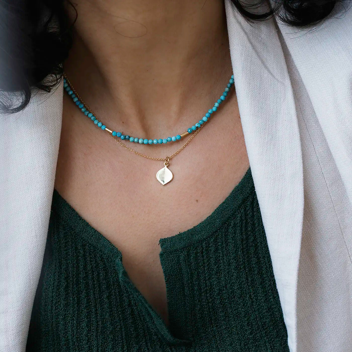 Eine Person trägt zwei Halsketten: eine mit kleinen, runden, türkisfarbenen Perlen und eine zarte goldene Kette mit einem glänzenden, blattförmigen Anhänger. Die Person trägt ein dunkelgrünes Oberteil mit Struktur und einen hellen Blazer darüber. Das Bild zeigt den Hals- und oberen Brustbereich, das Gesicht ist nicht sichtbar. Die Haare der Person sind dunkel und lockig.