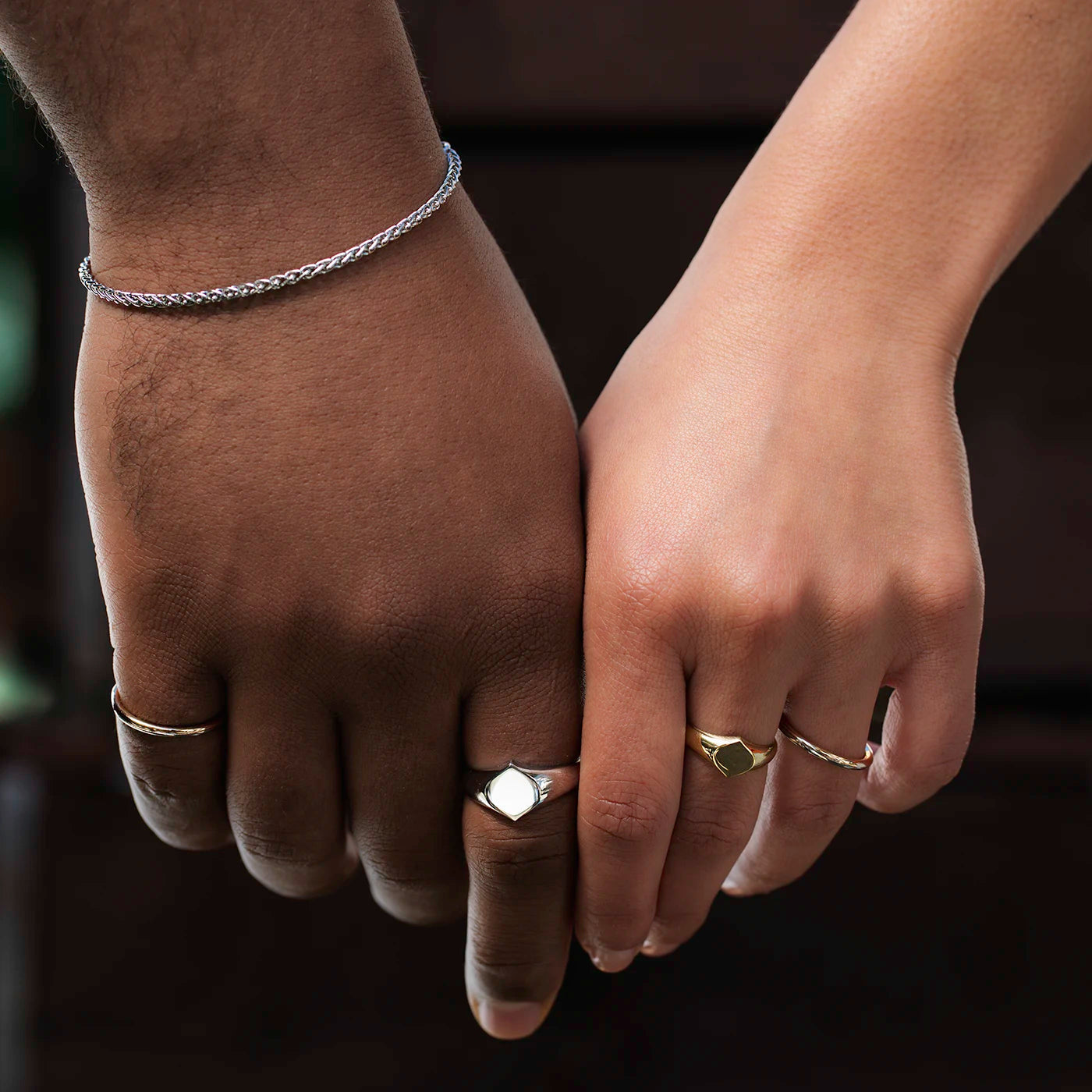 Zwei Hände, eine dunklere und eine hellere, halten sich. Beide tragen mehrere Ringe, eine Hand zudem ein geflochtenes Armband aus Platin.