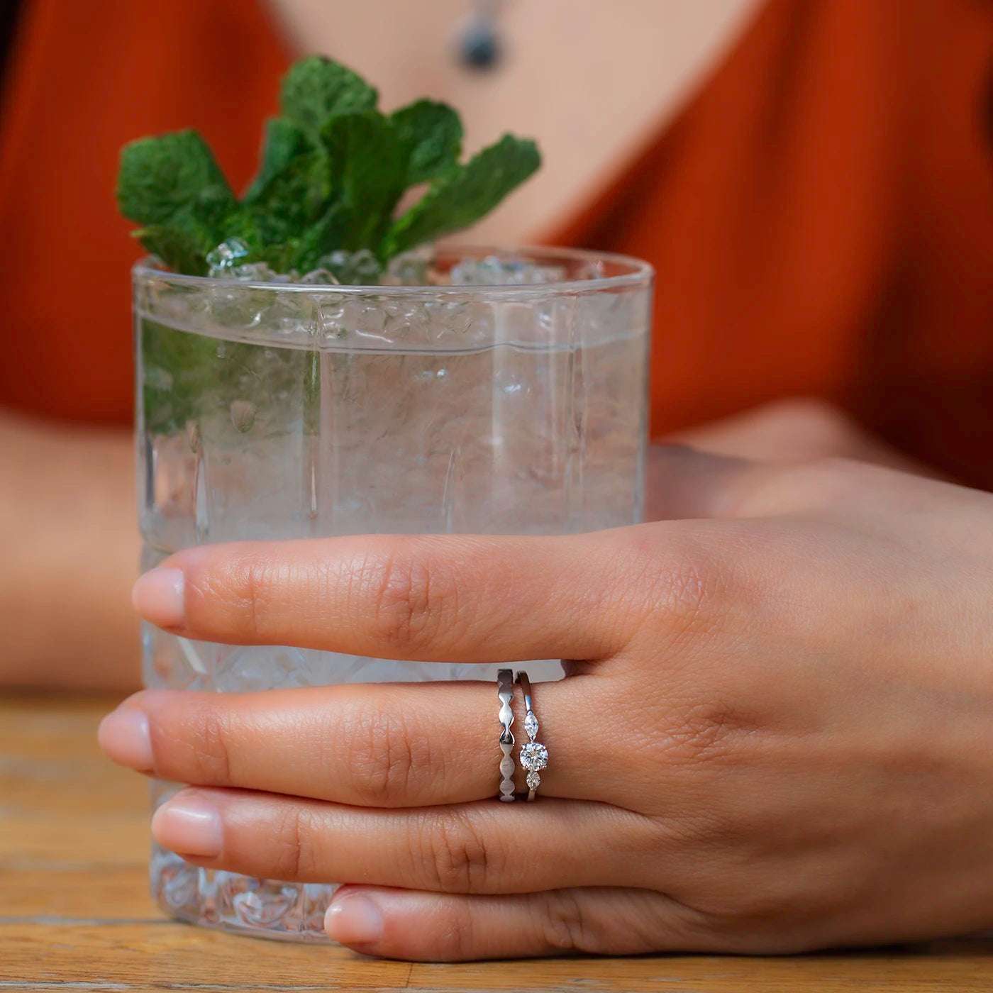 Ring Wave Platin die Ringschiene ist geschwungen wie Wellen Frontansicht an Modelhand Mittelfinger mit Zusteckring Petite Fleur in Platin Hand haelt Glas closeup