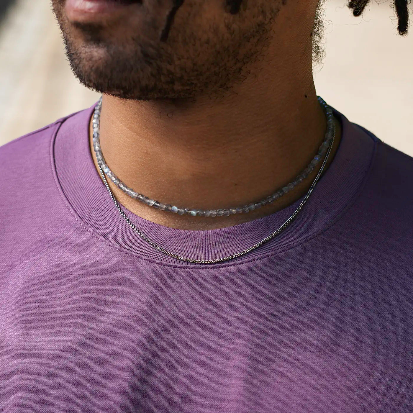 Ein Mann trägt ein lilafarbenes T-Shirt. Um seinen Hals liegen zwei Ketten: eine besteht aus kleinen, glänzenden Labradorit-Perlen, die nah beieinander aufgereiht sind und einen leichten Schimmer aufweisen; die andere Kette darunter hat eine feine, silberfarbene Struktur und wirkt wie ein schmales Platinelement. Das Bild konzentriert sich auf den Hals- und Schulterbereich, das Gesicht ist nur teilweise zu sehen. Im Hintergrund ist ein unscharfer, heller Bereich erkennbar.