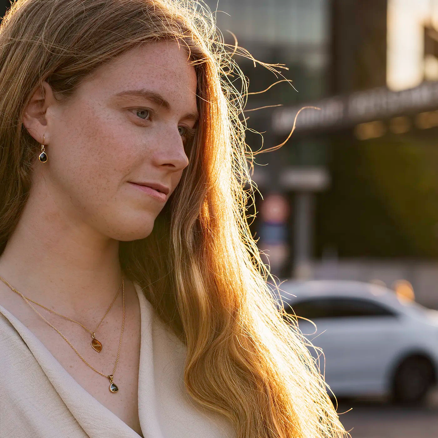 Eine Frau mit langen, blonden Haaren ist im Profil zu sehen. Sie trägt einen hellen Oberteil, goldene Ohrhänger mit dunklem Tropfenanhänger, sowie zwei goldene Halsketten, eine davon mit einem kleinen Blattanhänger und die andere mit einem dunklen, tropfenförmigen Anhänger.