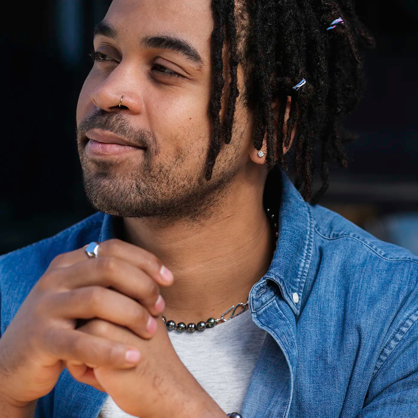 Ein junger Mann mit dunklem Teint, kurzen Dreadlocks mit kleinen Perlen, Nasenpiercing und Ohrring, trägt Jeanshemd, graues T-Shirt, Halskette mit Kugeln sowie einen Ring am Finger, Hände verschränkt, schaut zur Seite und wirkt entspannt.