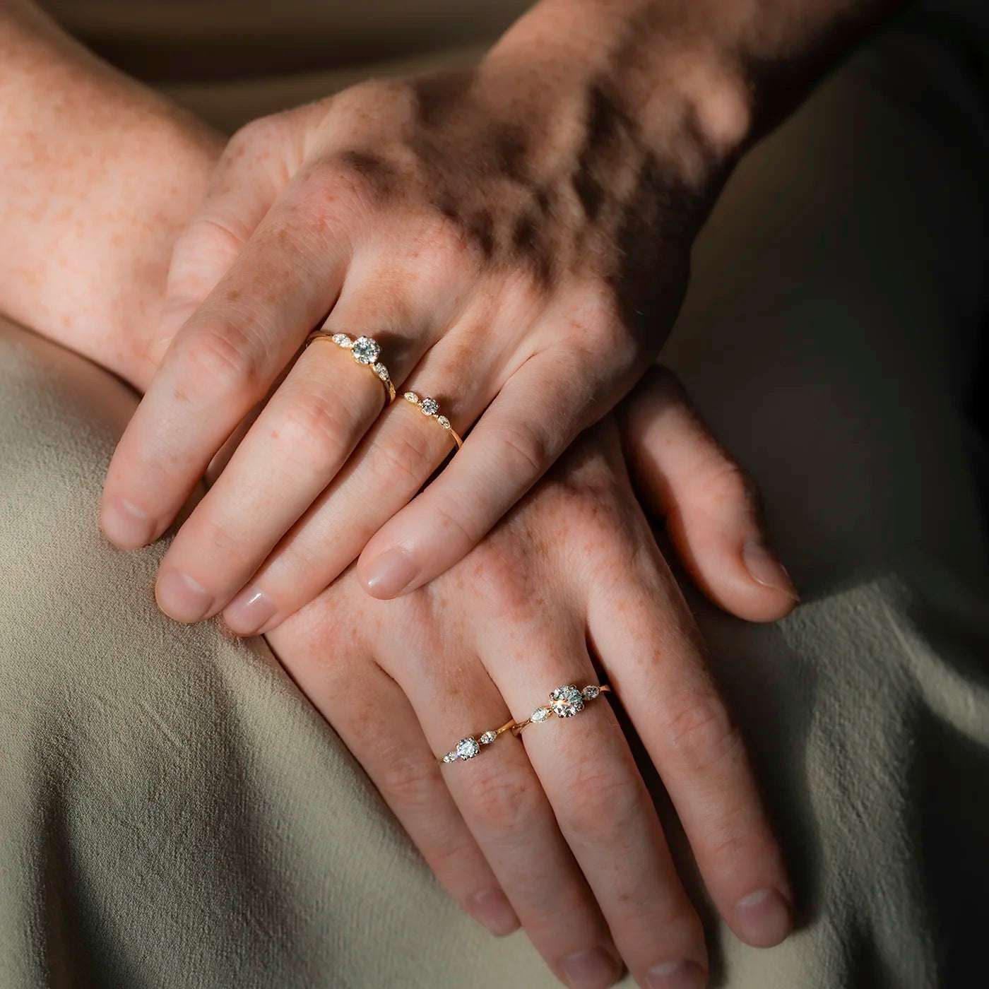 Ringe Petite Fleur Mittelstein 0.50ct und 0.75 Brillant je von 2 Navette Diamanten flankiert Gelbgold in floralem Design an Modelhand Ringfinger und Mittelfinger auf Schoss