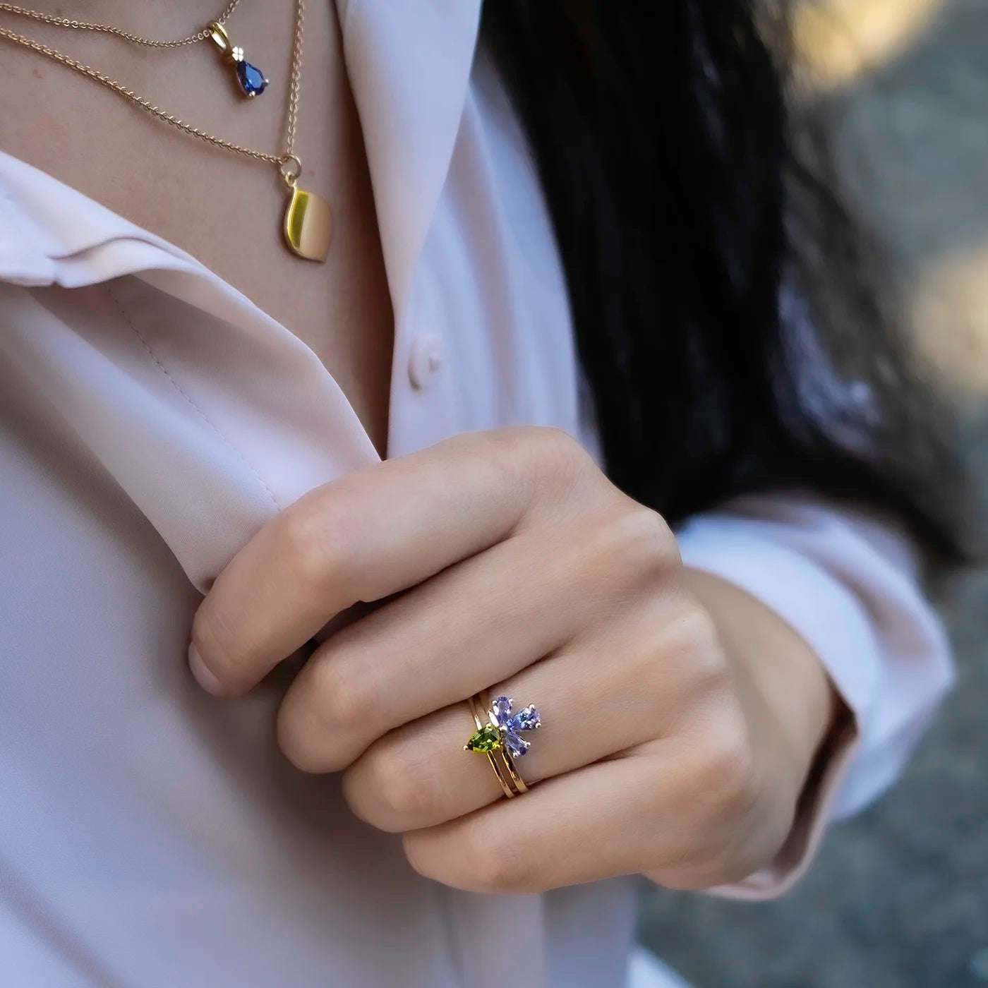 Eine Person trägt eine helle Bluse mit offenem Kragen. Am Hals befinden sich zwei goldene Ketten: Eine mit einem goldenen Anhänger, die andere mit einem dunkelblauen, tropfenförmigen Steinanhänger. Die Hand zeigt drei filigrane Goldringe, einer davon mit einem großen, hellvioletten, blütenförmigen Stein, der zweite mit einem kleinen grünen Stein verziert. Im Hintergrund ist das Haar der Person dunkel und leicht gewellt.