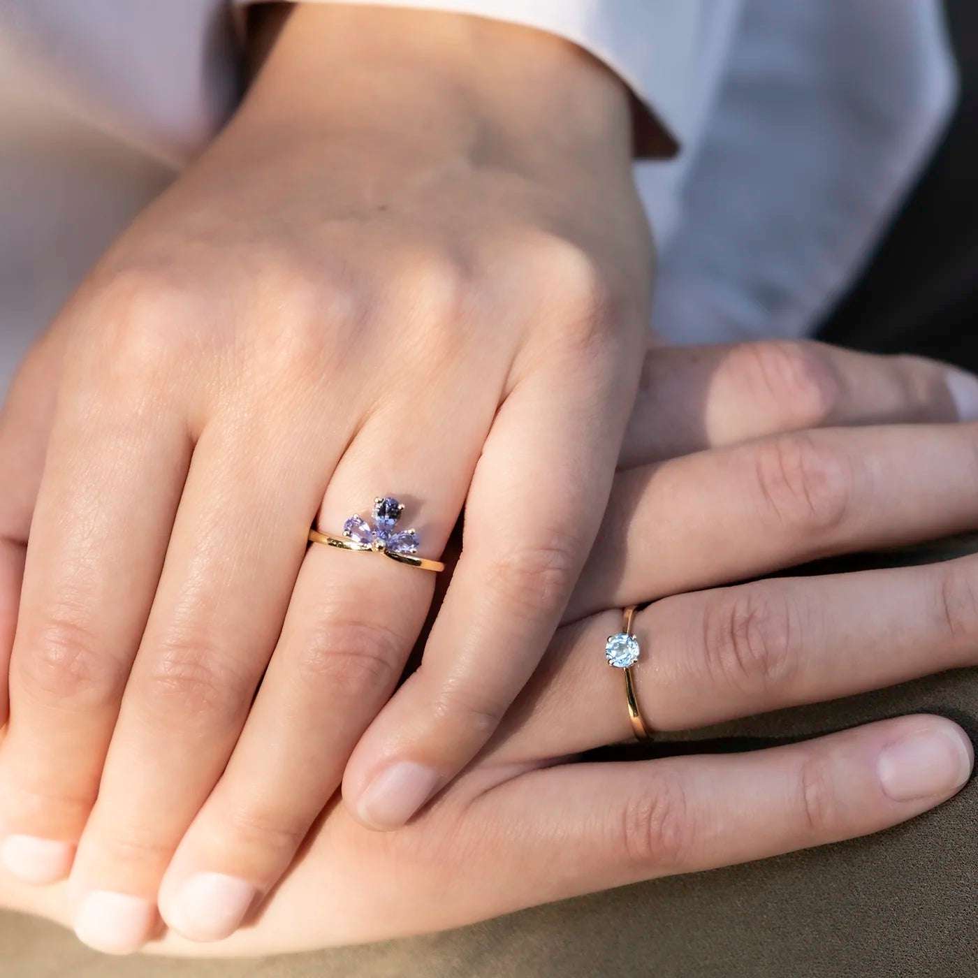 Zwei Hände liegen übereinander, beide tragen Ringe aus Gold. Einer der Ringe hat eine Blütenform mit lila Steinen, der andere einen einzelnen Stein. Hauttöne sind hell, der Hintergrund ist unscharf. Die Personen tragen helle Kleidung.