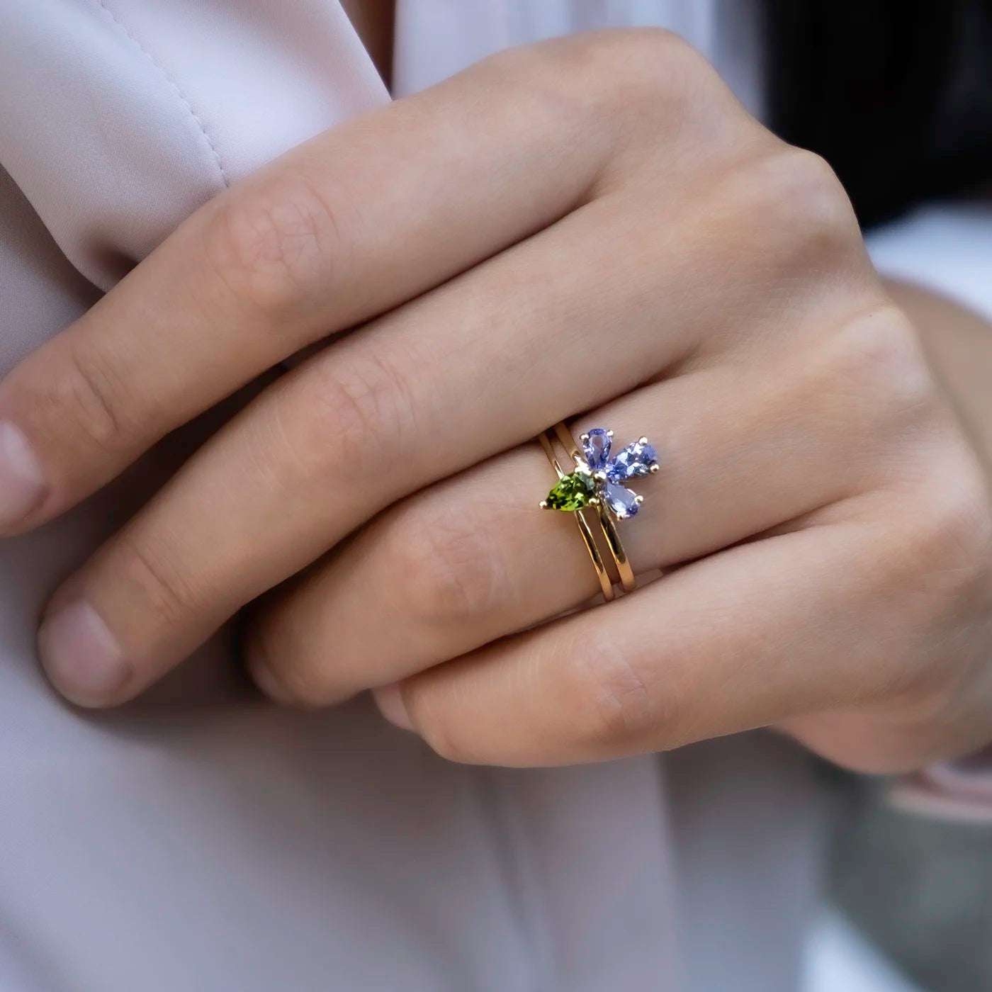 Eine Hand mit hell lackierten Nägeln, die einen goldenen Ring mit blütenförmigen, hellblauen und grünen Edelsteinen trägt. Die Ringe sind übereinander am Ringfinger getragen, der Hintergrund zeigt eine helle Kleidung.