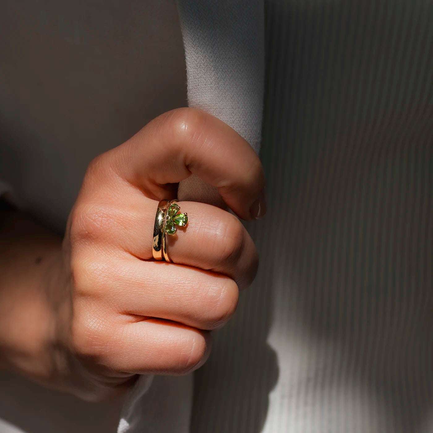 Eine Hand mit hellem Teint hält einen weißen Stoff. Am Finger trägt sie zwei Ringe: Ein glänzender, breiter Roségoldring und ein Zusteckring mit grünem, blütenförmigem Turmalin. Der Hintergrund ist weich und dezent, mit sanften Schatten. Die Ringe stehen im Mittelpunkt, besonders der grüne Turmalin, der durch Licht reflektiert wird. Die Hand ist entspannt und gepflegt, die Struktur des Stoffes wirkt fein und leicht gerippt.