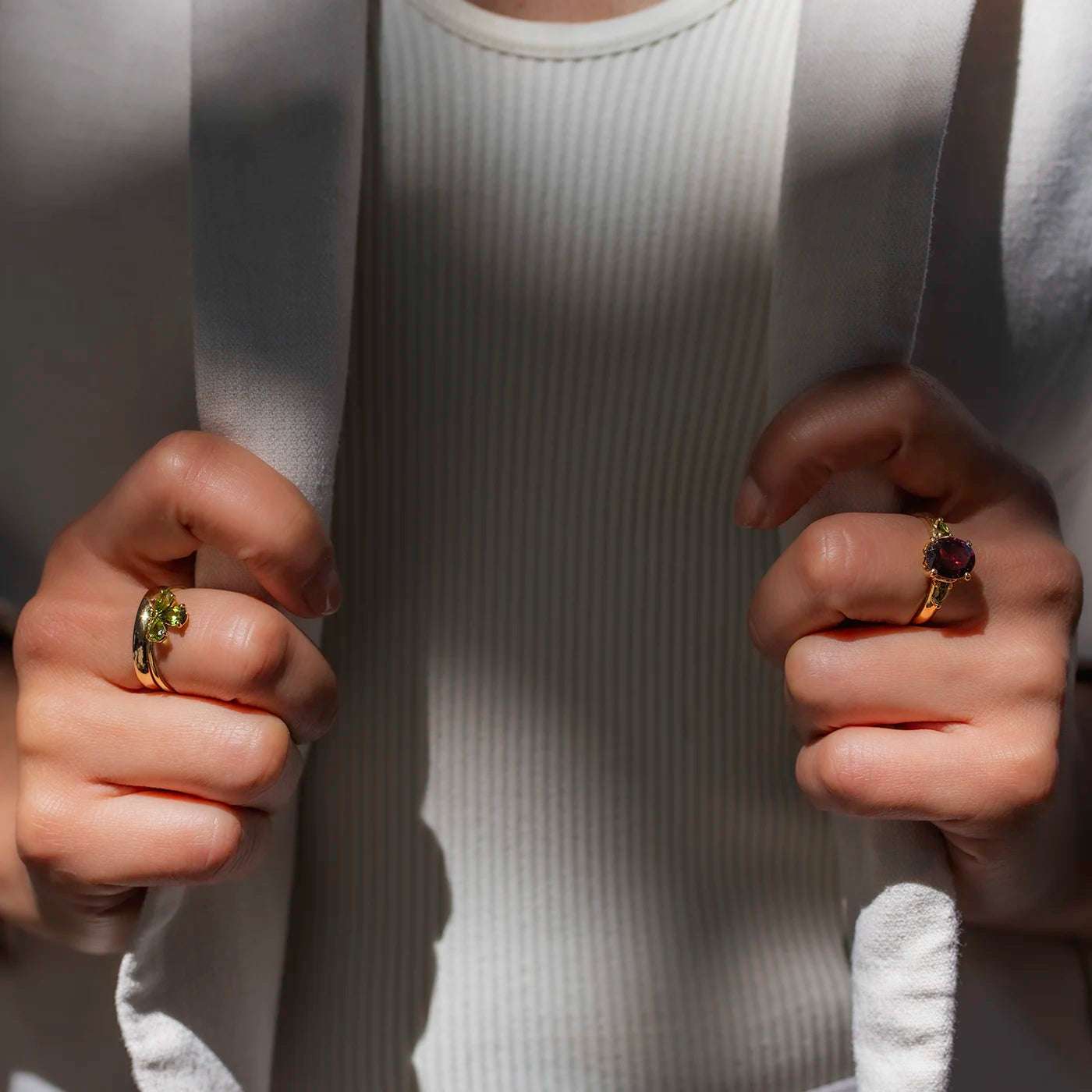 Eine Person trägt einen weißen Cardigan über einem weißen, gerippten Oberteil. An beiden Händen werden goldene Ringe mit auffälligen, runden Edelsteinen getragen, einer grün und einer rot. Die Person hält die Seiten des Cardigans mit den Händen fest. Licht und Schatten fallen auf die Szene.