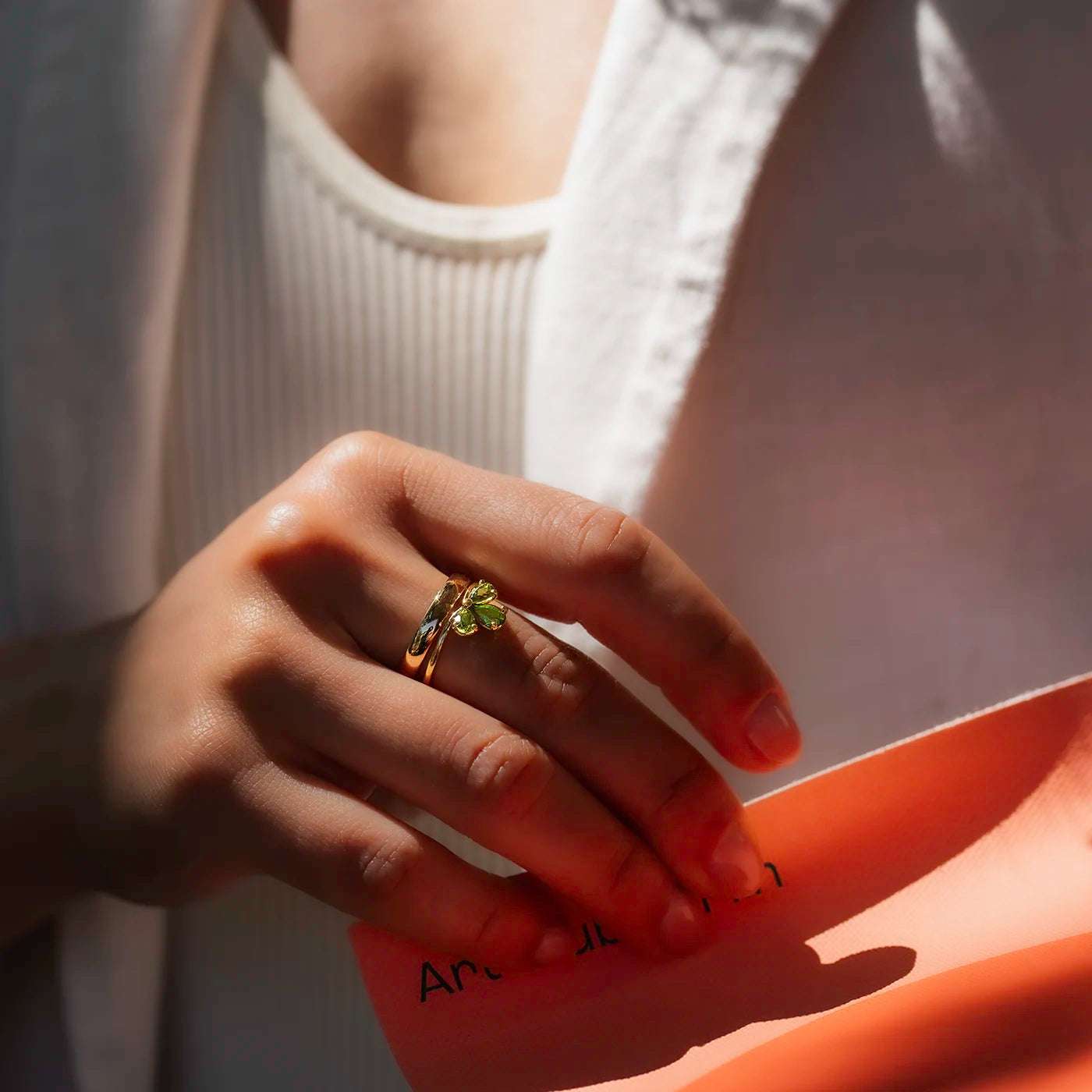 Eine Hand mit einem goldenen Ring mit grünem, blütenförmigem Stein hält ein orangenes Blatt Papier. Die Person trägt ein weißes, geripptes Oberteil und eine helle Jacke. Das Licht fällt sanft auf die Hand und betont den Schmuck.