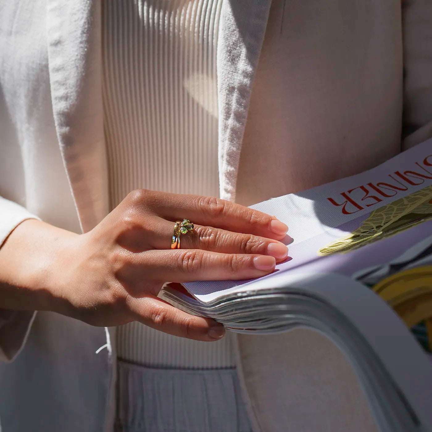Eine Hand mit elegant lackierten Fingern hält eine Zeitschrift. Die Person trägt einen goldenen Ring mit grünem Stein, ein heller Blazer und ein cremefarbenes, geripptes Oberteil sind sichtbar. Das Licht fällt freundlich auf die Szene, Details eines goldenen Motivs und roter Schrift auf dem Magazin sind erkennbar.