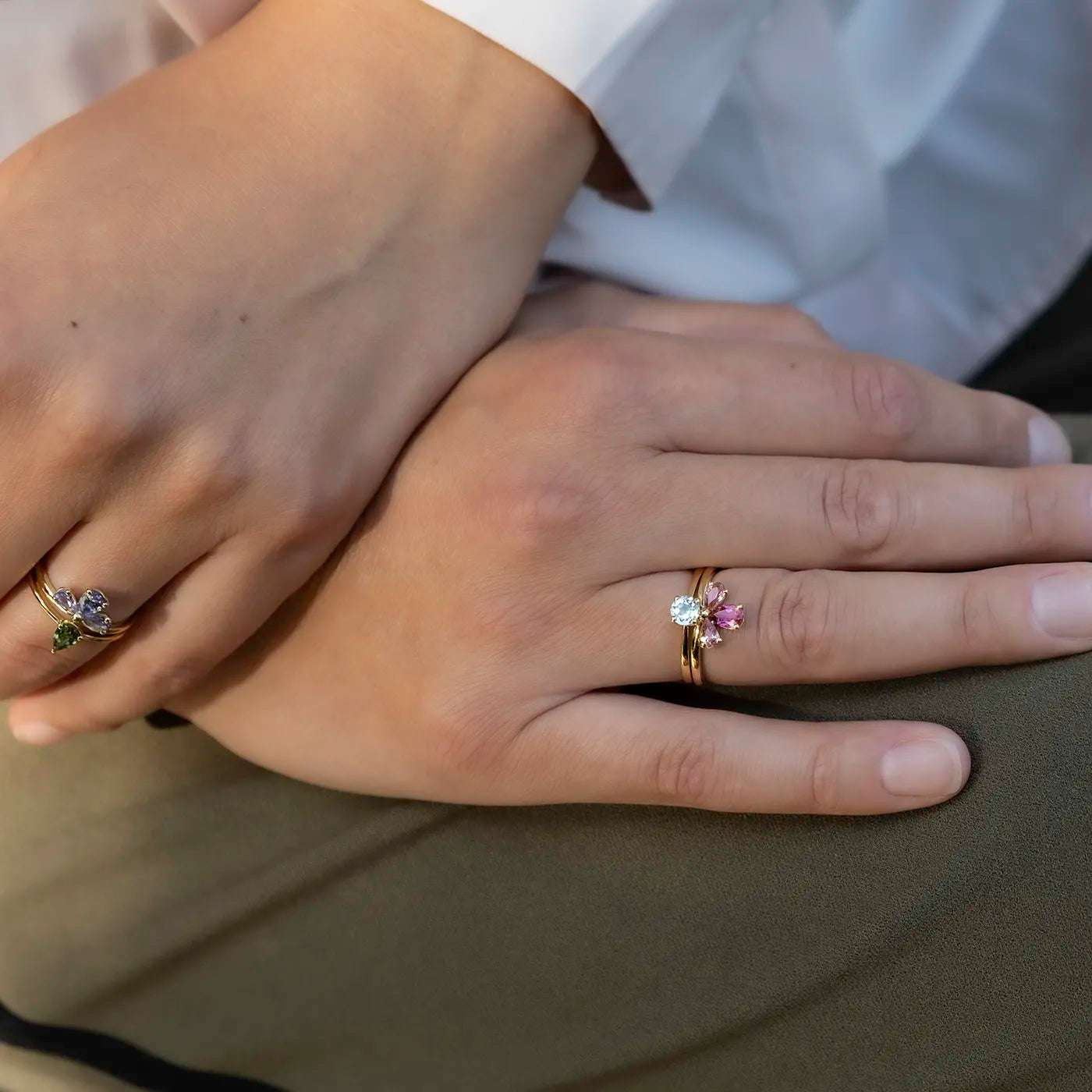 Zwei Hände mit mehreren goldenen Ringen, verziert mit verschiedenfarbigen, blütenförmig angeordneten Edelsteinen, darunter Rosa und Grün.