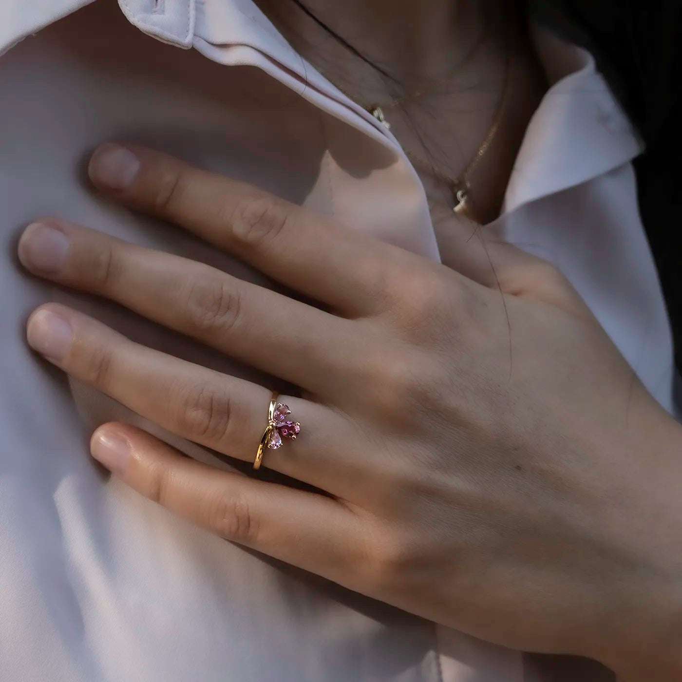 Eine Hand mit schlanken Fingern liegt auf einer weißen Bluse. Am Ringfinger trägt die Person einen goldfarbenen Ring mit mehreren rosa Steinen, die wie eine Blüte angeordnet sind. Die Haut ist hell, die Bluse leicht geöffnet, und es ist eine zarte Halskette mit kleinem Anhänger am Hals zu sehen. Das Licht ist weich, der Fokus liegt auf dem Ring.