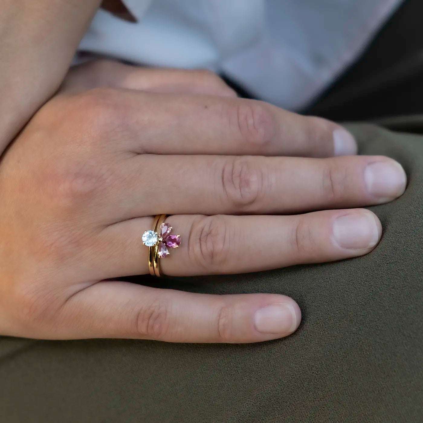 Eine Hand mit natürlichem Hautton liegt auf einem Khaki-Stoff. Am Ringfinger werden zwei goldene Ringe getragen. Einer der Ringe ist mit drei auffälligen Edelsteinen besetzt: einem weißen, einem rosafarbenen und einem kleineren hellrosa Stein. Die Steine sind blütenförmig arrangiert. Die Fingernägel sind kurz und ungepflegt. Im Hintergrund erkennt man verschwommen weiße und dunkle Kleidung.