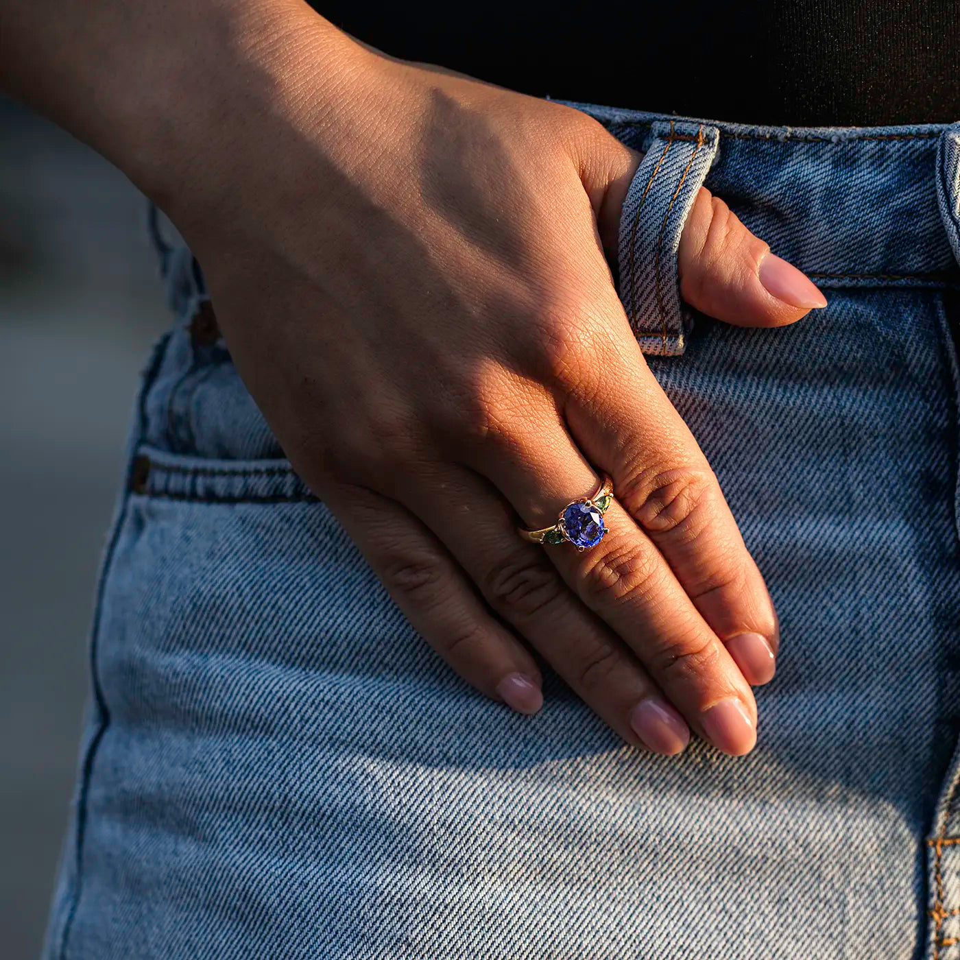 Eine Hand mit lackierten Fingernägeln liegt auf einer blauen Jeans. Am Ringfinger wird ein auffälliger goldener Ring mit einem großen, tiefblauen bis violetten Edelstein getragen. Die Hand hält sich lässig am Hosenbund fest.