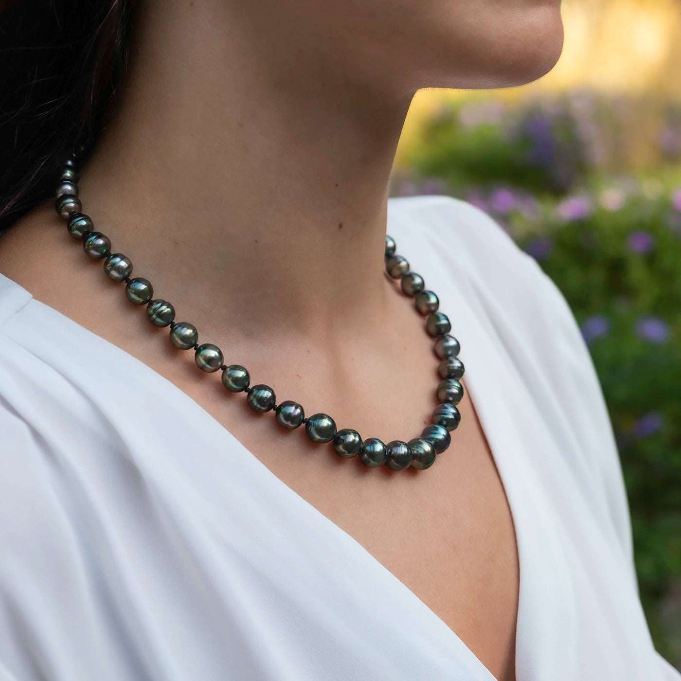 Kette aus Tahitiperlen mit Gelbgold Infinity Verschluss an Modelhals vor gruenen Blumen Closeup