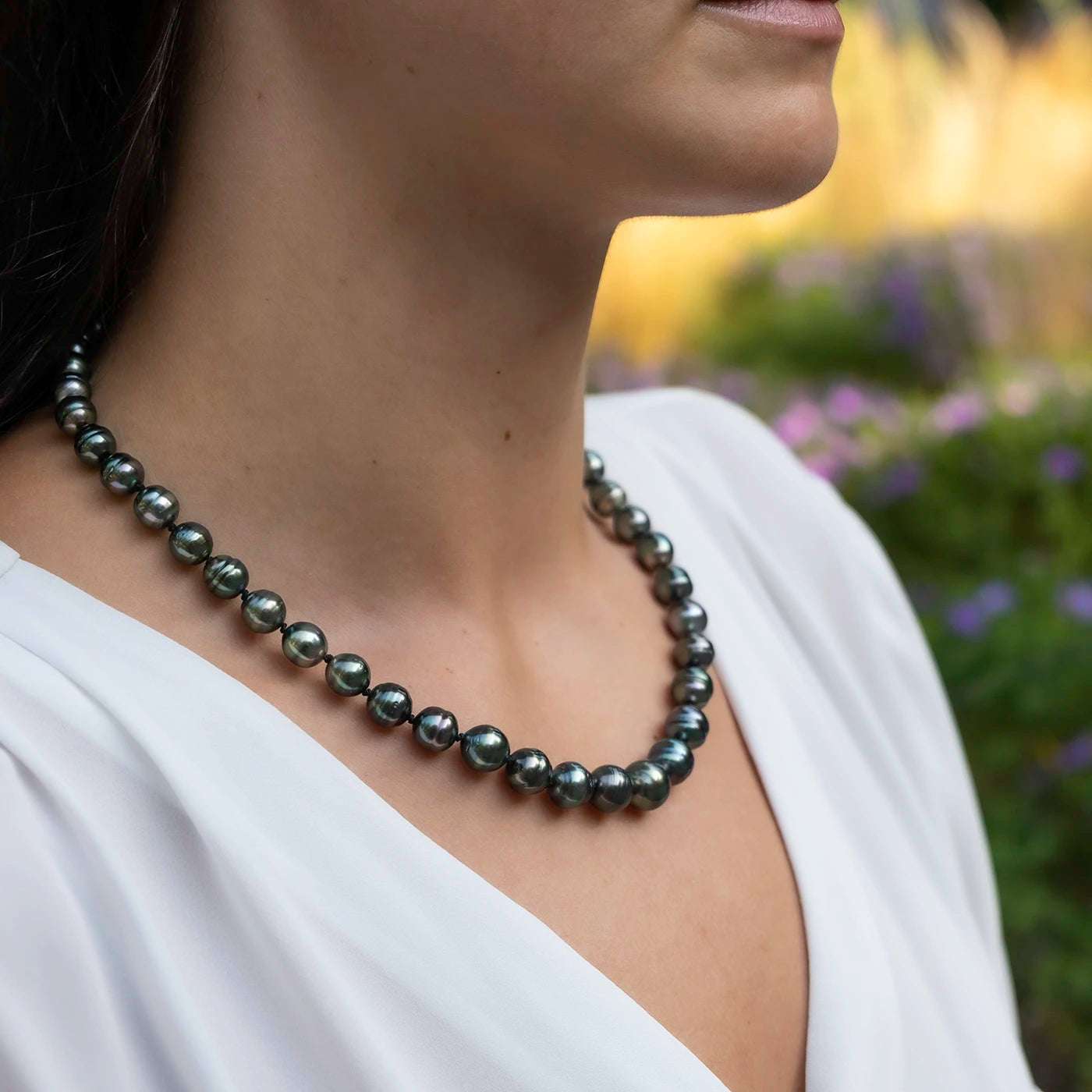 Kette aus Tahitiperlen mit Gelbgold Infinity Verschluss an Modelhals vor gruenen Blumen Closeup andere Haltung