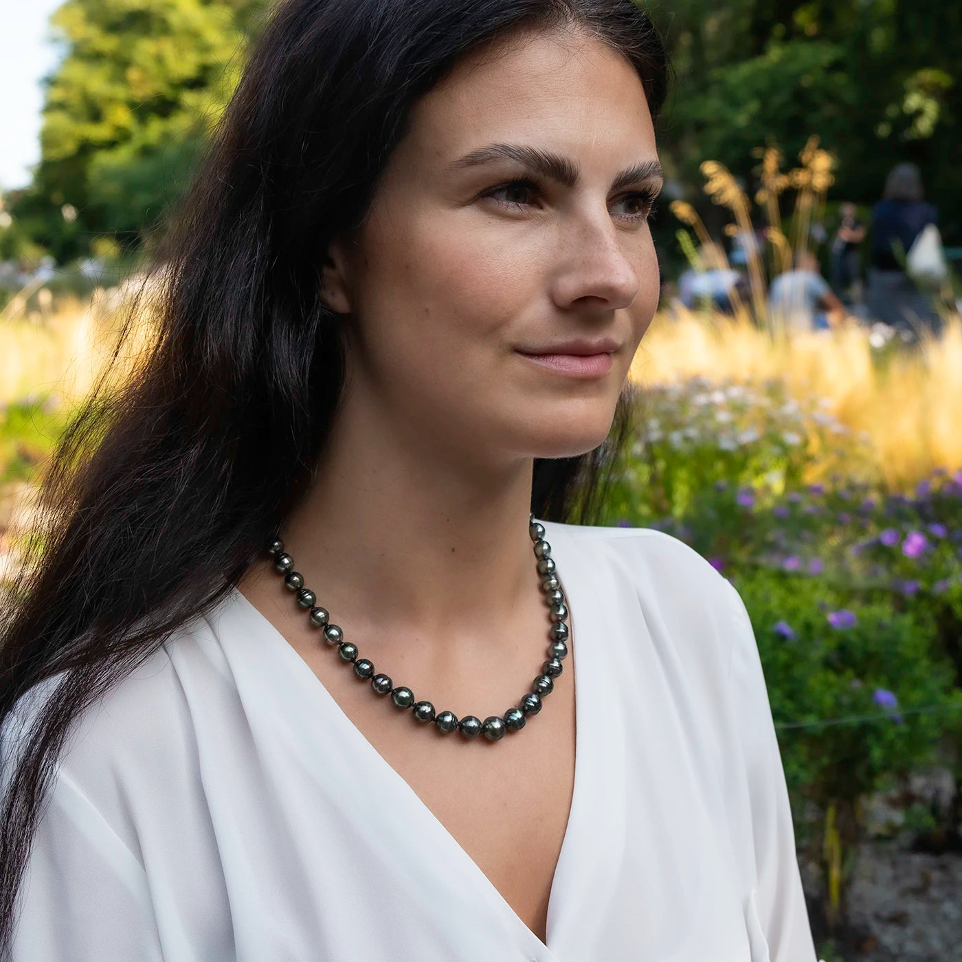 Kette aus Tahitiperlen mit Gelbgold Infinity Verschluss an Modelhals vor gruenen Blumen halber Oberkörper drauf