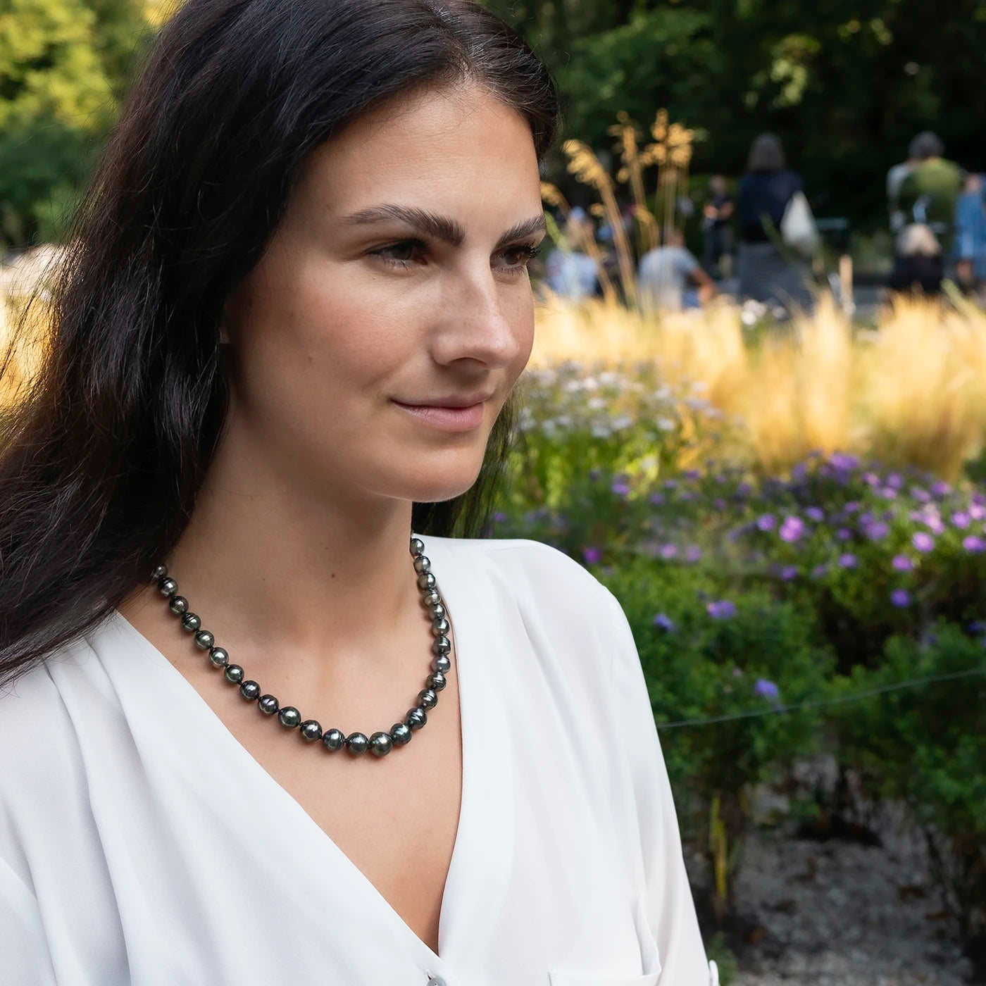 Kette aus Tahitiperlen mit Gelbgold Infinity Verschluss an Modelhals vor gruenen Blumen halber Oberkörper drauf weiterer Winkel