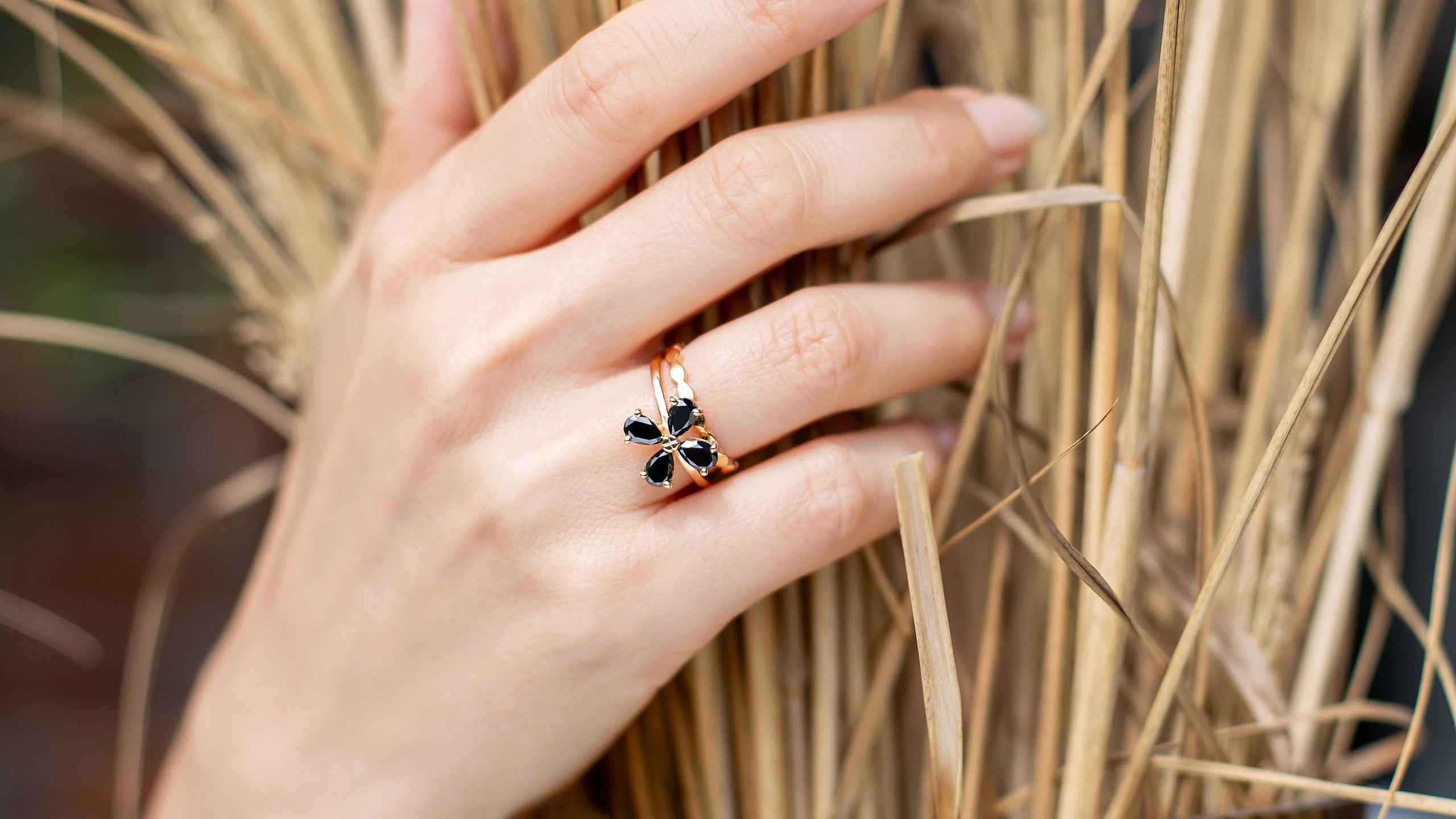 Ring Forever Blossom aus 750er Gold mit vier schwarzen Diamanten in klarer Blütenform, an der Hand getragen vor getrocknetem Ziergras