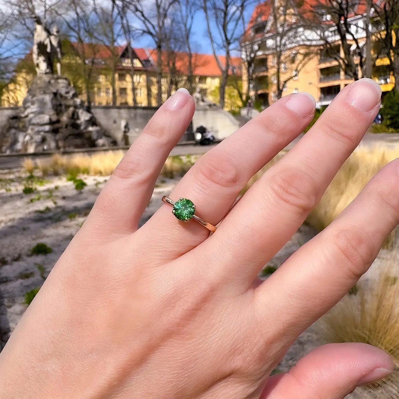 Petite Fleur Solitär Ring aus Roségold mit grünem Turmalin, getragen am Ringfinger vor Berliner Altbaufassaden und Brunnen