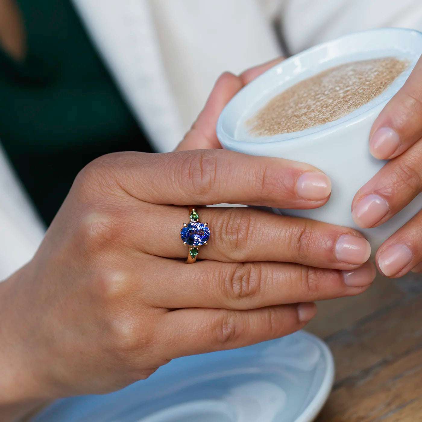 Ring Fleur Unikat blauer Tansanit und zwei gruene Turmaline Gelbgold  im floralem Design an Modelhand haelt Kaffeetasse closeup