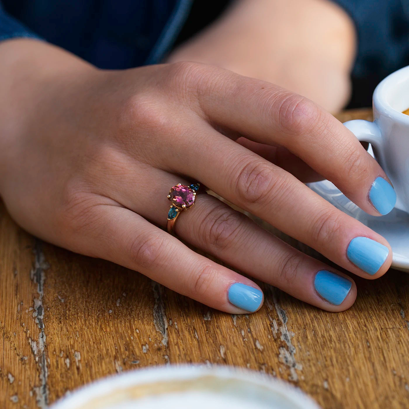 Ring Fleur Unikat roter Turmalin und zwei blaue Topas im floralem Design an Ringfinger Model Hand auf Kaffeetisch closeup