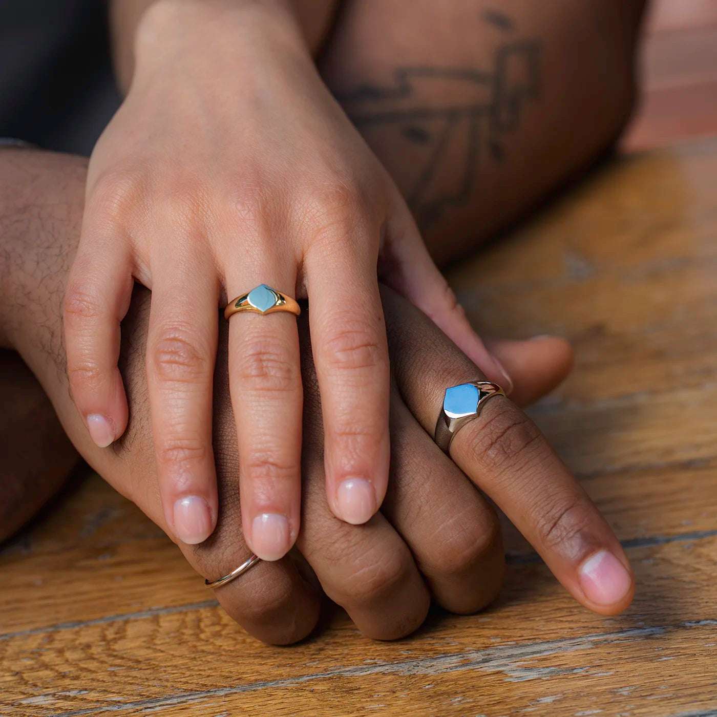 Siegelring Damen Gelbgold und Herrenring Platin mit grosser Flaeche in Form Josefine Logo fuer Wunschgravur Frontansicht an weiblich und maennlicher Modelhand Ringfingern Haende uebereinander gelegt auf Tisch